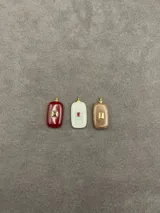 Dije de piedra natural con forma rectangular y bordes redondeados, con detalle central de engaste metálico dorado y piedra pequeña incrustada. Disponible en tres variantes de color: rojo, blanco traslúcido y marrón.