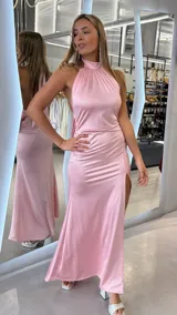 Vestido largo rosado de satén, con escote halter alto y espalda descubierta. Presenta un tajo pronunciado en la pierna izquierda y diseño drapeado en los laterales.