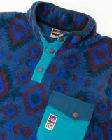 Buzo de polar fleece con diseño estampado geométrico en tonos azules, bordó y verde. Presenta cuello alto con cierre de botones a presión, bolsillo frontal con solapa en contraste y puños elásticos.