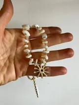 Gargantilla artesanal compuesta por piedras naturales blancas irregulares intercaladas con pequeñas cuentas doradas. Presenta un dije central en forma de sol de bronce y cuenta con cadena de alargue con cierre de mosquetón.