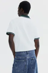 Polo de manga corta en piqué de algodón, color blanco con cuello y bordes de mangas en color verde oscuro con detalles de líneas blancas. Presenta tapeta con botones.