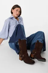 Bota de caña media confeccionada en cuero gamuzado color chocolate. Presenta un diseño de estilo western con taco cuadrado de 7 cm y una correa decorativa en el tobillo con herrajes metálicos plateados.
