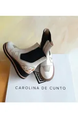 Botas cortas Carolina De Cunto modelo Kiria, confeccionadas en cuero de color blanco con detalles pintados a mano. Poseen inserciones elásticas laterales en color negro, suela de goma con plataforma y detalles texturizados en el talón.