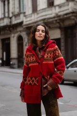 Saco tejido a mano con diseño jacquard en tonos rojo, naranja y marrón. Presenta cuello tipo polo, cierre frontal con botones grandes, mangas largas y tajos laterales en el ruedo.