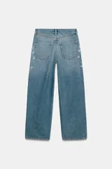 Jeans tiro medio de corte relaxed leg y largo completo, confeccionados en denim azul claro. Presentan detalles de aplicaciones de pedrería y joyas a contraste en los laterales superiores y bolsillos delanteros, estilo clásico de cinco bolsillos y cierre frontal con cremallera y botón metálico.