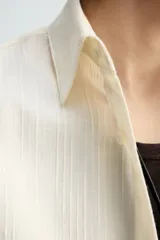 Camisa de algodón color crema o blanco roto, de corte regular fit, con cuello solapa, manga larga con puño abotonado y bolsillo plastrón en el pecho. La tela presenta una textura sutil de rayas verticales en relieve.