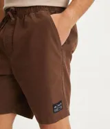 Bermuda masculina color marrón, confeccionada en tejido plano de algodón texturizado con poliéster. Presenta cintura elástica con cordón de ajuste y un tag de marca en el lateral inferior.