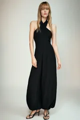 Mono largo de color negro con cuello halter cruzado y hombros descubiertos. Presenta un diseño de piernas tipo bombacho con bajos ajustables mediante lazadas y cierre lateral oculto.