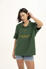 Remera de manga corta color verde oscuro, estilo polo con cuello y abertura frontal. Presenta un bordado central con el texto "PulauCrew" y un laurel en la parte superior.