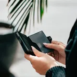 Billetera delgada de cuero sintético color gris oscuro/negro, con mecanismo de eyección para tarjetas y cierre a presión. El nombre "BALEINE" está grabado en la parte inferior frontal.