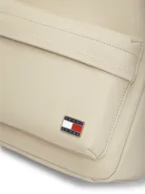 Mochila Tommy Jeans de diseño minimalista en color beige, confeccionada en poliuretano de acabado liso. Cuenta con un compartimento principal con cierre, un bolsillo frontal con cremallera, asa superior y correas ajustables. Incluye el logo metálico de la marca en la parte inferior frontal.