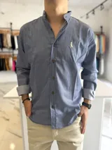 Camisa de manga larga azul con rayas verticales blancas, cuello mao y cierre frontal con botones oscuros. Presenta un pequeño bordado en el pecho.