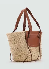Bolso tipo cesta de fibras naturales con solapa y asas de cuero vacuno en color marrón. Presenta un diseño tejido a mano con detalles de costuras decorativas en las asas y tiras finas colgantes.