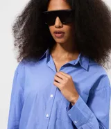 Camisa alargada de manga larga, confeccionada en tricolina de algodón con diseño de rayas finas azules y blancas. Cuenta con cuello inglés, cierre frontal con botones y un bolsillo aplicado en el pecho.