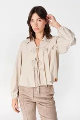 Blusa de algodón color beige con cuello camisero, mangas largas abullonadas con puños elásticos y cierre frontal mediante tres lazos para anudar.