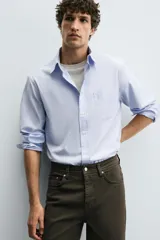 Camisa de algodón de corte regular, color celeste claro con finas rayas blancas verticales. Presenta cuello abotonado (button-down), manga larga con puños abotonados y un bolsillo plastrón en el pecho izquierdo.