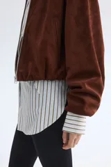 Campera corta marrón de tejido suave con terminación cepillada, cuello camisero, hombros caídos y cierre frontal. Cuenta con bolsillos laterales ribeteados y elástico revestido en puños y dobladillo.