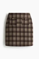 Pollera de tiro alto a la rodilla confeccionada en punto bondeado con diseño a cuadros en tonos marrón y beige. Presenta dos bolsillos de parche frontales con botones metálicos dorados, cierre oculto posterior y bajo recto.