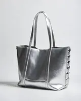 Cartera tipo tote de símil cuero plateado con doble asa de hombro, detalles de tachas en los laterales y placa metálica con logo de la marca en el frente.
