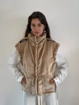 Chaleco de peluche color beige con cuello alto, cierre frontal, bolsillos laterales y detalles de etiquetas en los hombros.