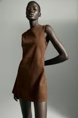 Vestido corto de cuello barco y manga sisa, confeccionado en tejido efecto ante. Presenta un diseño en línea evasé con detalle de pliegues laterales y cierre de cremallera oculta en la espalda.