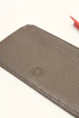 Monedero rectangular de cuero vacuno con textura granulada en color marrón topo. Cuenta con cierre superior metálico y un tirador decorativo de cuero en color naranja con un nudo. Presenta un pequeño logo grabado en el centro.