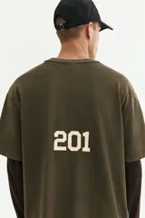 Remera de algodón de punto pesado con efecto lavado, color verde militar. Presenta un corte boxy (holgado) con cuello redondo ribeteado y un pequeño estampado blanco en el pecho con texto y logos.