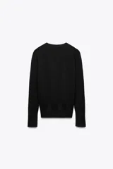 Jersey de punto liso color negro, con cuello redondo y manga larga. Presenta una hilera de cuatro botones dorados decorativos en el puño de cada manga.