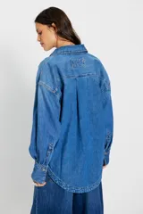 Camisa de denim azul de calce amplio, con cuello clásico, botonadura frontal, bolsillo plaqué en el pecho, hombros caídos y puños anchos abotonados.