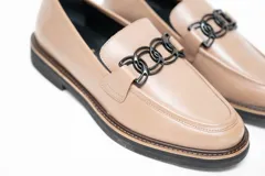 Mocasín de cuero vacuno color visón, con diseño clásico de punta redondeada y costuras visibles. Presenta un adorno metálico negro en el empeine y suela plana de goma negra.