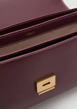 Bolso tipo bandolera de tamaño pequeño con acabado efecto piel en color bordó. Presenta una solapa frontal con cierre metálico a presión, costuras visibles en contraste y una correa larga ajustable con herrajes dorados.