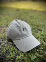 Gorra tipo dad hat en color gris lavado, con diseño de dos hojas bordadas en hilo negro en la parte frontal. Cuenta con visera curva y estructura flexible.