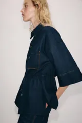 Camisa de denim azul oscuro con cuello solapa, mangas amplias por debajo del codo y bolsillos de parche en el frente. Cuenta con cintura ajustable mediante cordón y cierre frontal con botones metálicos.