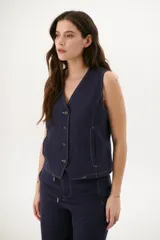 Chaleco sin mangas color crudo, con escote en V y cierre frontal de tres botones oscuros. Presenta costuras expuestas en contraste oscuro que definen el diseño y bolsillos delanteros simulados.