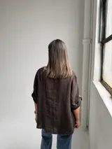 Blusa de lino color marrón chocolate, de corte holgado, con cuello redondo, mangas tres cuartos con trabilla y botón para sujetar remangadas, y detalle de alforzas verticales en el frente.