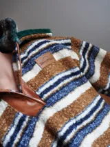 Buzo canguro oversize de paño de lana gris con capucha estampada y cuello de corderito blanco. Cuenta con aberturas laterales en el ruedo y detalles de cuero sintético marrón en el cierre frontal y etiqueta.