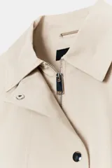 Trench corta de color beige, con cuello solapa, mangas largas con trabillas en los hombros, bolsillos delanteros de vivo y cierre frontal cruzado con botones a presión.
