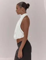 Top blanco con diseño halter, cuello alto y espalda descubierta. Presenta un frente cruzado con pliegues, bajo elástico y lazo de ajuste en la espalda.