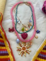 Set de tres collares con dijes de temática veraniega. El primero es de cuentas rosadas con dijes de sol, cangrejo y otros. El segundo es de cuentas verdes con un dije de corazón. El tercero es dorado con dijes varios.