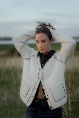 Saco tipo cardigan de punto color blanco con detalles de pespuntes decorativos en color negro a lo largo de los bordes y costuras. Presenta cuello en V, cierre frontal con botones y mangas largas.