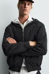 Cazadora relaxed fit color negro, confeccionada en tejido técnico ligero que repele el agua. Cuenta con cuello subido, capucha ajustable, manga larga y cierre frontal con cremallera. El interior está forrado con tejido de estructura abierta y tiene bolsillos frontales ocultos en la costura. Los acabados son elásticos.