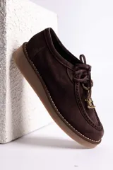 Zapato tipo mocasín acordonado de gamuza color marrón oscuro, con suela de goma color caramelo y detalle de dije metálico dorado en forma de bota colgando de los cordones.