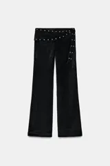 Pantalón de tiro alto con diseño tipo pareo en la cintura, ajustable con lazada y decorado con tachas metálicas. Presenta una silueta acampanada y cierre lateral con cremallera oculta.