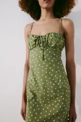 Vestido midi verde militar con estampado de lunares rosados, de corte recto y tirantes finos cruzados en la espalda. Presenta escote con frunces y detalle de lazo en el centro.