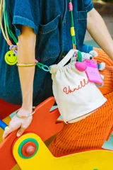 Llavero para bolso con cuerda trenzada en colores violeta y verde, decorado con un dije circular de acrílico rosa con diseño de carita feliz con ojos en forma de corazón. Incluye argollas metálicas de colores y una etiqueta pequeña de silicona.