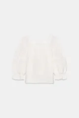 Blusa blanca de manga corta abullonada con puño elástico, escote cuadrado y detalles de puntillas y pinzas verticales en el frente.