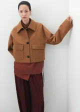 Chaqueta de corte recto color tostado, con cuello camisero, manga larga, dos bolsillos de parche con solapa en el frente y cierre frontal de botones oscuros.