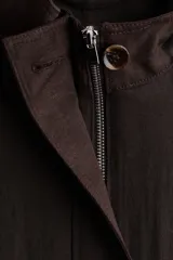 Campera corta oversize de color marrón, confeccionada en tejido de lino. Presenta cuello elevado, cierre frontal oculto con solapa y botones visibles, mangas raglán con puños abotonados, bolsillos delanteros con solapa y cordón ajustable en el bajo.