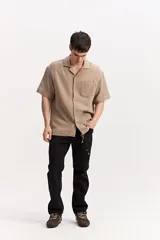Camisa de manga corta color beige confeccionada en muselina de algodón, con cuello abierto, cierre frontal de botones y un bolsillo aplicado en el pecho.