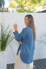 Camisa de jean de corte oversize, con cuello clásico, cierre frontal de botones y mangas largas. Presenta un diseño con bolsillos laterales integrados y ruedo redondeado.